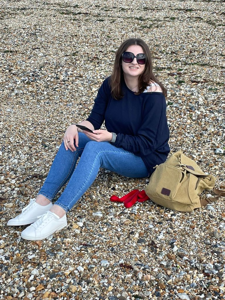 The author (long brown hair, sunglasses, blue jumper, blue jeans, white trainers) sitting on a pebble beach
